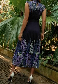 Robe florale sombre avec des manches volantes, présentant des motifs violets et verts, une jupe fluide, portée avec des chaussures à talons blancs.