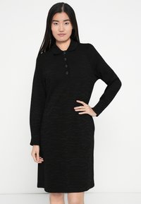 Robe noire avec col polo, manches longues, patte de boutonnage à cinq boutons et motif subtil en texture ondulée. Longueur midi et coupe décontractée.