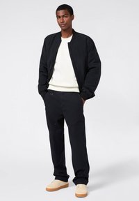 Giacca bomber nera sopra un felpa color crema, abbinata a pantaloni neri e sneakers beige chiaro. Tessuto morbido e vestibilità rilassata in tutto.