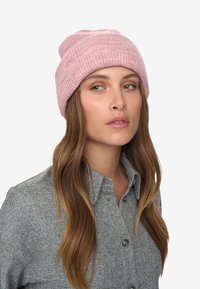 Frau mit langen braunen Haaren, die eine pinkfarbene Strickmütze und ein graues Hemd mit Knöpfen trägt, und leicht nach rechts schaut, vor schlichtem Hintergrund.
