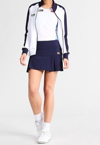 Falda de tenis plisada azul marino combinada con una blusa blanca y una chaqueta con cremallera con acentos en azul marino; incluye zapatillas blancas y calcetines tobilleros.