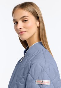 Jasnoniebieska pikowana kurtka bomberka z kołnierzem typu stand, srebrnym zamkiem błyskawicznym i naszywką z logo na lewym rękawie. Gładka faktura i wykończenia w kontrastowym kolorze.