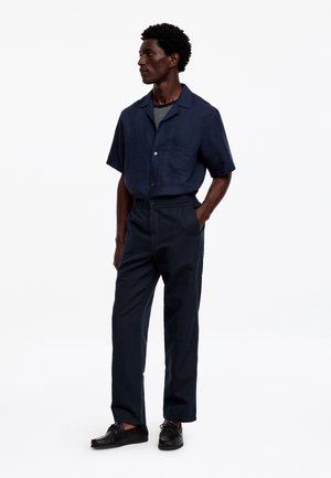 Homme portant une chemise bleu marine à manches courtes boutonnée, un sous-vêtement rayé, un pantalon sombre et des chaussures noires, debout avec une main dans la poche.