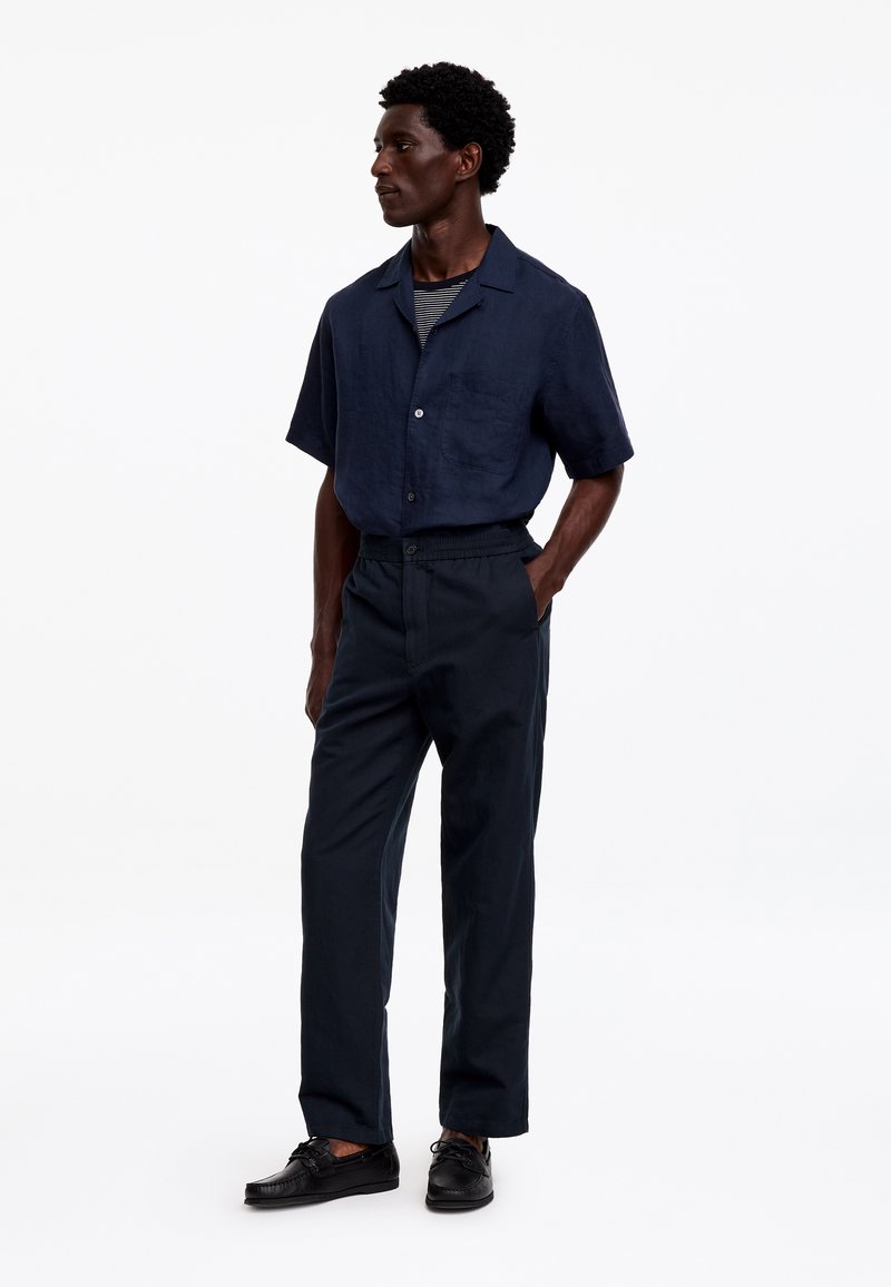 Homme portant une chemise bleu marine à manches courtes boutonnée, un sous-vêtement rayé, un pantalon sombre et des chaussures noires, debout avec une main dans la poche.