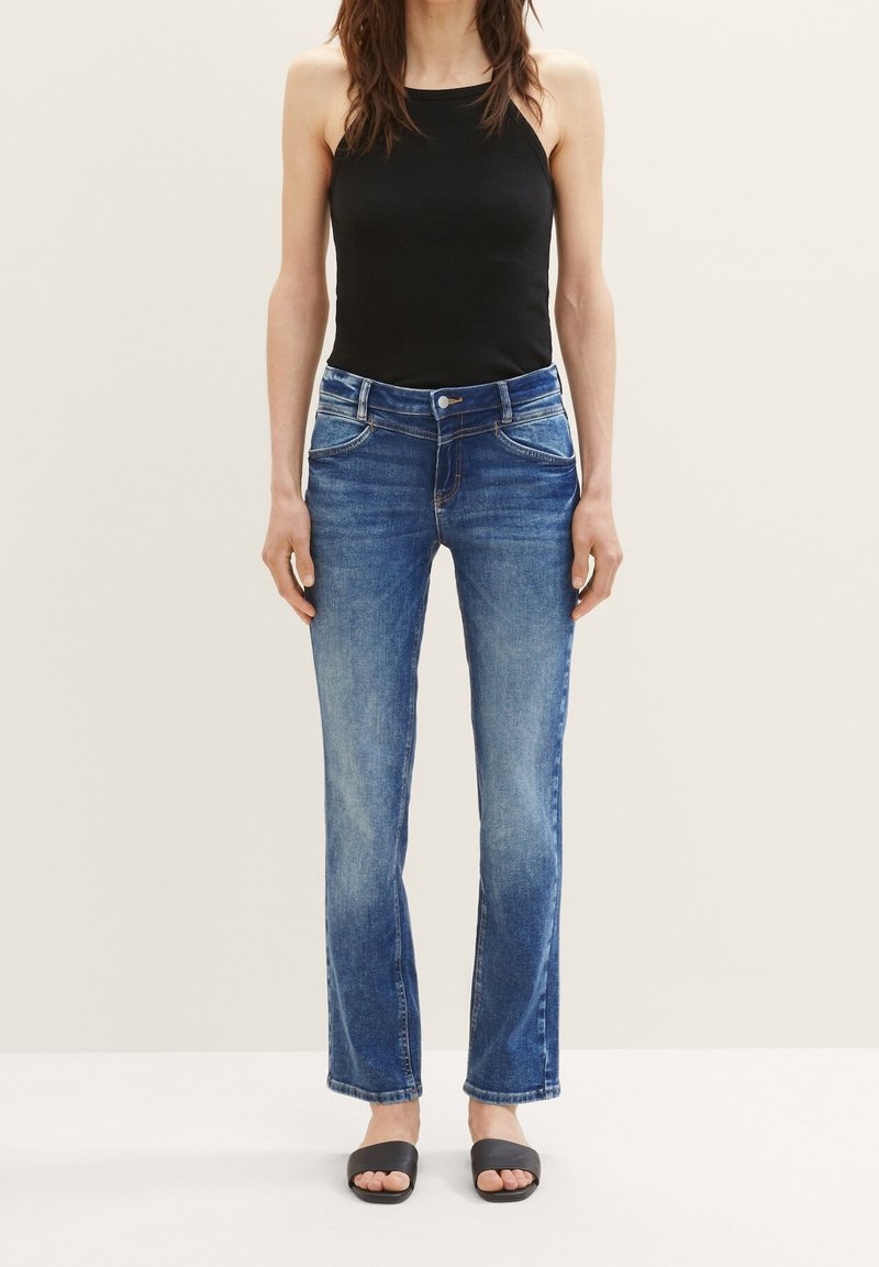 Jean en denim bleu à coupe droite, avec cinq poches, texture délavée et fermeture par bouton. Associé à un débardeur noir et des sandales noires.
