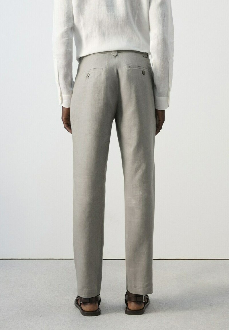 Personne portant un pantalon gris clair sur mesure et une chemise blanche à manches longues, vue de dos, debout sur un sol clair.