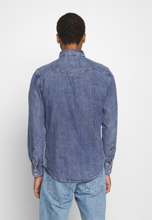 Homme portant une chemise en denim bleue à manches longues et un jean bleu clair, debout dos à la caméra sur un fond blanc uni.