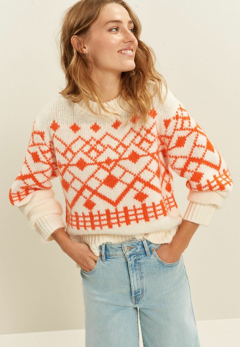 Weißer Strickpullover mit orangefarbenen geometrischen Mustern, gerippten Bündchen und Saum, kombiniert mit hellblauen hoch geschnittenen Jeans.