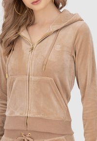 Felpa con zip in velluto beige, dotata di tasca frontale, zip dorata e logo ricamato. Tessuto morbido. Ambientazione in studio con sfondo neutro.