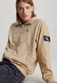 Chaqueta beige con cremallera y cuello alto, que presenta detalles del logo negro 'cK' y puños elásticos. Tela suave y ligera, ideal para superponer.