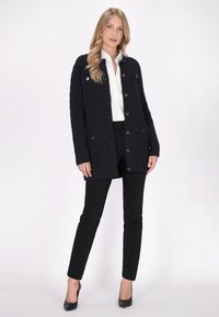 Cardigan lavorato a maglia blu marina con motivo a trecce, chiusura a bottoni, tasche sul petto e un taglio rilassato, abbinato a pantaloni neri slim e tacchi alti.