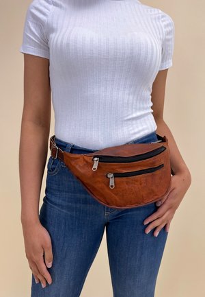 Vrouw die een wit geribd T-shirt met korte mouwen, een blauwe spijkerbroek en een bruine leren heuptas met twee ritsvakken draagt.