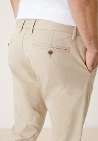 Homme portant un chino beige avec une poche arrière boutonnée et les mains dans les poches avant, vêtu d'une chemise blanche sur un fond uni.