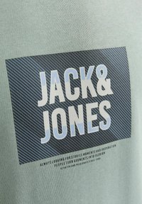 Camiseta verde claro con un gráfico negro que incluye "JACK & JONES" en letras grandes, blancas y azules; tela texturizada con un sutil patrón.