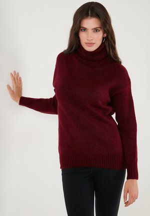Woman with long brown hair wearing hoop earrings, a burgundy turtleneck sweater, and black pants, leaning against a white wall.