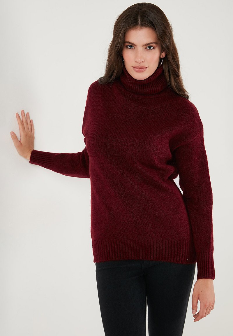 Femme aux longs cheveux bruns portant des boucles d'oreilles créoles, un pull col roulé bordeaux et un pantalon noir, appuyée contre un mur blanc.