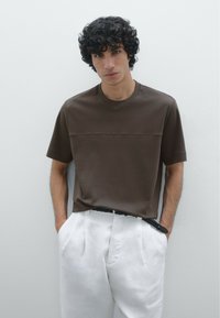 T-shirt a maglia marrone con maniche corte, caratterizzato da una cucitura orizzontale sul petto, abbinato a pantaloni bianchi plissettati e una cintura nera.
