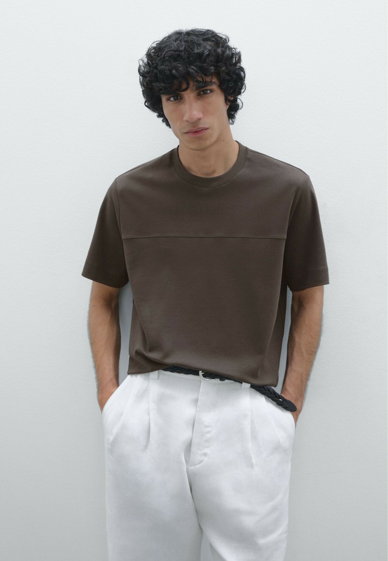 T-shirt a maglia marrone con maniche corte, caratterizzato da una cucitura orizzontale sul petto, abbinato a pantaloni bianchi plissettati e una cintura nera.