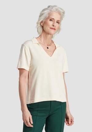 Kremfarbener Poloshirt mit kurzen Ärmeln, V-Ausschnitt und geripptem Kragen. Weicher Stoff, lockere Passform, kombiniert mit dunkelgrünen Anzughosen.
