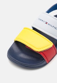 Kindersandaal met marineblauwe zool, rode en gele voorbanden, en witte enkelband met het merk "Tommy Hilfiger" en logo op een witte achtergrond.
