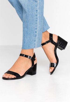 Voeten met zwarte blokhak sandalen van lakleer met enkelbandjes, gecombineerd met lichtblauwe cropped spijkerbroeken.