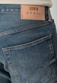 Jeans de mezclilla azules con una etiqueta de cuero beige que lleva el texto "EDWIN", bolsillos traseros con detalles de costura curva y un acabado desgastado.