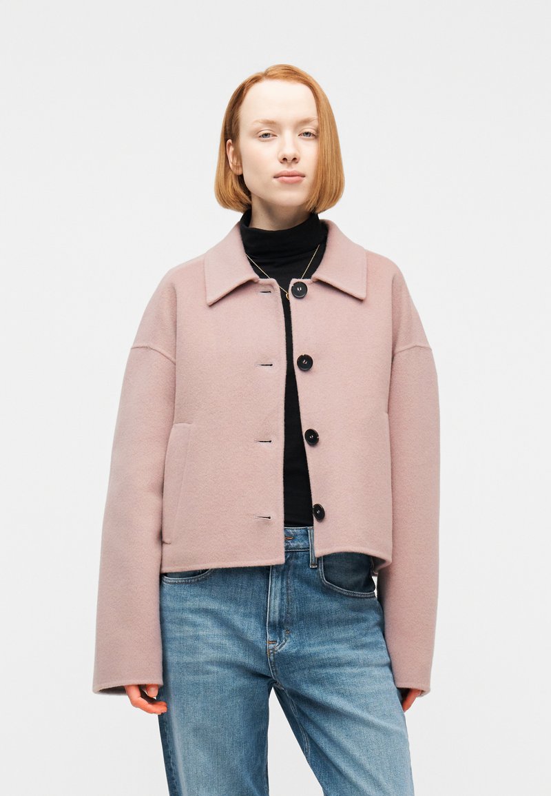 Νεαρό άτομο με κοντά κόκκινα μαλλι ά φοράει παλτό ανοιχτού ροζ χρώματος με κουμπιά πάνω από μαύρο ζιβάγκο και μπλε τζιν, με λευκό φόντο.