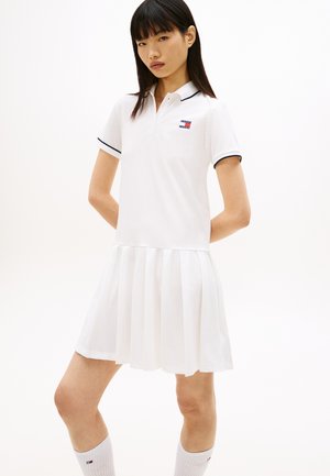 Giovane donna che indossa un vestito polo bianco con gonna plissettata e logo sul petto, in piedi con le mani dietro la schiena contro uno sfondo semplice.