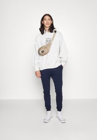 Riñonera beige llevada sobre un sudadera blanca con un logo, acompañada de pantalones de chándal azul marino y zapatillas altas blancas. Diseño simple y ajuste casual.