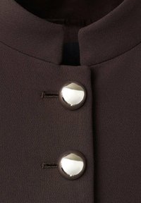 Tela de chaqueta marrón con cuello redondo, dos botones plateados brillantes y sutiles acentos de costura cerca de los botones. Textura suave.