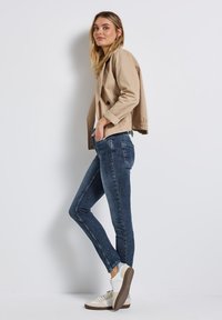 Veste cropped beige avec deux boutons, jean kaki ajusté, et baskets blanches. La veste a des poignets élastiques et des poches latérales.