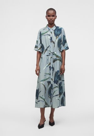 Femme portant une robe midi bleu clair avec des imprimés floraux foncés, manches courtes, col, taille nouée et talons noirs pointus.