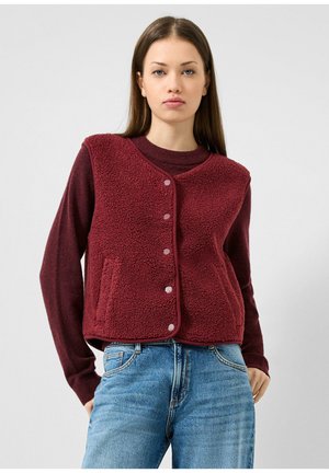 Jeune femme portant un gilet texturé bordeaux sur un haut à manches longues et un jean bleu, debout devant un fond clair uni.