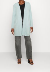 Helles blaues Cardigan mit offenem Vorderteil, langen Ärmeln und Taschen, kombiniert mit einem schwarzen Oberteil und grauen, hochtaillierten Jeans, getragen mit schwarzen hohen Schuhen.