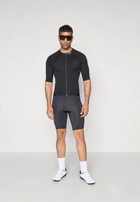 Sort cykeltrøje med korte ærmer og lynlås foran, kombineret med mørkegrå shorts. Modellen bærer solbriller og hvide sportssko.