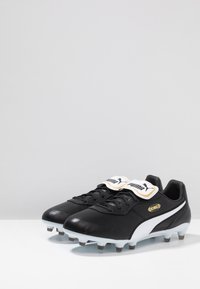Botas de fútbol de cuero negro con acentos blancos, superficie texturizada, logo dorado y tacos cónicos para tracción. Cuello acolchado para mayor comodidad.