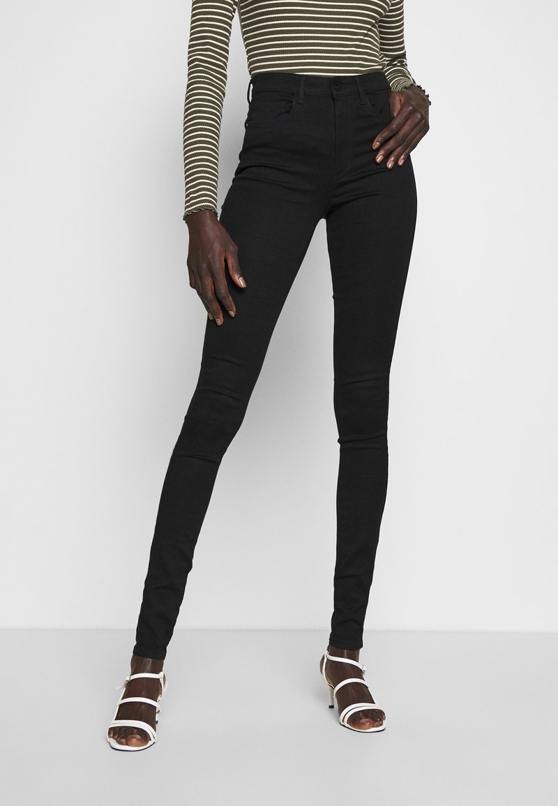 Personne portant un jean skinny noir taille haute, un haut à manches longues rayé, et des sandales blanches à talons avec des brides, debout devant un fond blanc.