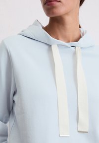 Hellblaues Sweatshirt mit Kapuze, ausgestattet mit weißen Kordeln und silbernen Metallenden. Weicher Stoff und minimales Design.