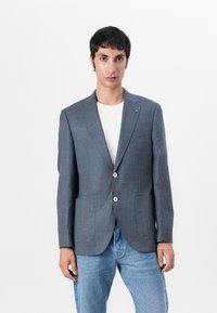 Blazer i blå rutmönster med två knappar, slagkrage och sidofickor, bärs över en vit T-shirt och blåjeans.