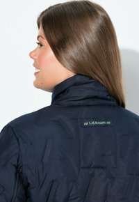 Marinblå vadderad jacka med hög krage, textil med strukturerat mönster och en logotags i grönt på baksidan.
