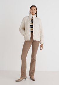 Quiltet beige jakke med høj krave, tætsiddende design og sidelommer, parret med stribet top og brune flared bukser.