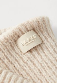 Strikket beige lue med ribbestruktur, utstyrt med et kremfarget lærmerke gravert med "BARTS AMSTERDAM" for merkevaren.
