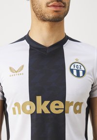 Jersey a righe bianche e blu navy con scollatura a V, motivi geometrici, testo dorato "nokera", logo "CASTORE" e un distintivo circolare della squadra.
