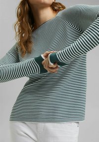 Femme portant un t-shirt à manches longues rayé vert et blanc avec des trous pour les pouces, ajustant les manches devant un fond gris uni.