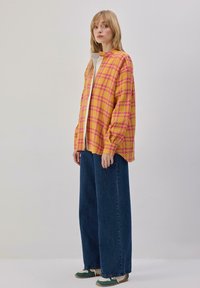 Veste-chemise à manches longues à carreaux jaunes et roses, portée sur un haut blanc et associée à un jean bleu à jambes larges et des baskets vertes.