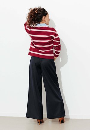 Femme debout contre un mur blanc, une main dans un pantalon noir à jambes larges, portant des talons marron et une chemise rouge à rayures à manches longues, les cheveux tirés en arrière.