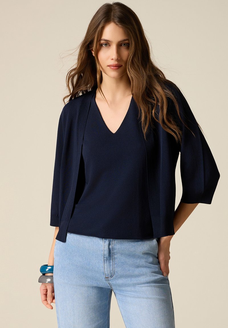 Donna con capelli lunghi e mossi indossa una maglia a scollo a V blu navy e un cardigan abbinato con jeans azzurri, in piedi davanti a uno sfondo beige.