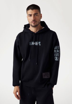 Schwarzer Hoodie aus weichem Baumwollmaterial mit einem grafischen Druck auf der Vorderseite, Kordelzügen und Text an den Ärmeln. Mit einem Logopatch an der unteren Seite.