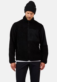 Czarna polarowa kurtka z kieszeniami na zamek, teksturowany materiał, wysoki kołnierz oraz gładkie czarne panele. Noszona ze dopasowaną dzianą czapką.