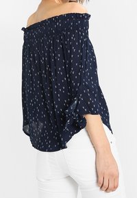 Donna che indossa una blusa blu scuro scollata con piccole stampe floreali bianche e pantaloni bianchi, rivolta di lato con un braccio visibile.